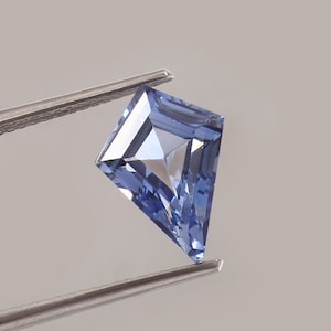 May include: A light blue, kite-shaped gemstone is held by tweezers. The gem's facets catch the light, creating a sparkling effect. The background is a soft, neutral grey. The gemstone is a kite cut.