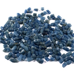 May include: A pile of small, rough-cut blue gemstones. The stones are various shades of blue, from light to dark, and have a slightly irregular, crystalline appearance. They are scattered on a white background.