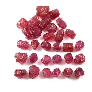 Puede incluir: Una colección de 27 piedras preciosas de rubí rojo en bruto. Los rubíes son de varios tamaños y formas, algunos tienen una forma más rectangular y otros son más redondeados.