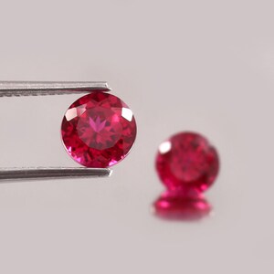 Puede incluir: Dos gemas redondas, facetadas, de color rojo intenso. Una es sostenida por unas pinzas plateadas, la otra está ligeramente desenfocada. Las gemas tienen una superficie brillante y reflectante. El fondo es gris neutro.