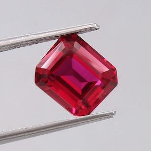 May include: A vibrant, deep red gemstone with an emerald cut, featuring sharp, clean facets that reflect light. The stone is held by a pair of silver-toned metal tweezers, showcasing its transparent, saturated color and rectangular geometric shape.