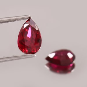 May include: Two faceted, pear-shaped rubies. One is held by tweezers, displaying a deep red hue with internal reflections. The other ruby is out of focus, resting on a reflective surface, also showing a deep red color.