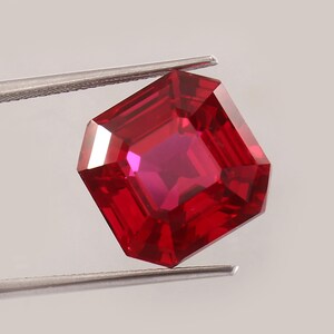 May include: A faceted, radiant-cut ruby gemstone held by tweezers. The gem is a deep, saturated red with multiple facets that reflect light. The shape is a modified square with beveled corners, showcasing the stone's brilliance.