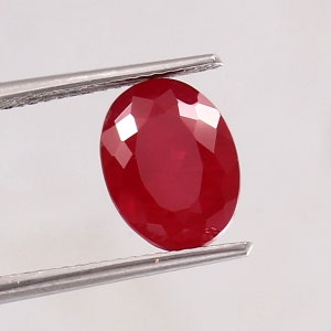 Könnte beinhalten: Ein leuchtend tiefroter, oval geschliffener Edelstein mit zahlreichen Facetten, die das Licht reflektieren. Der Edelstein wird sicher von einer silberfarbenen Metallpinzette gehalten und zeigt seine klare, polierte Oberfläche und satte Farbe.