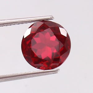 May include: A round, faceted gemstone with a deep, vibrant red color. The stone features a symmetrical, multi-faceted cut that reflects light across its surface, creating a brilliant appearance. It is held securely by silver-toned metal tweezers.