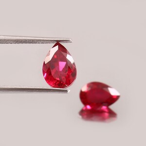Peut inclure: Deux rubis en forme de poire, facettés. L'un est tenu par une pince, l'autre repose sur une surface réfléchissante. Les pierres précieuses sont d'un rouge vif avec des reflets internes, mettant en valeur leur clarté et leur taille. Le fond est de ton neutre.
