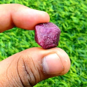 以下が含まれることがあります： 2 本の指で持たれた、深紅色のルビーの宝石。ルビーは自然な質感で、緑の草を背景にしています。宝石は、豊かな色合いと自然な質感を持っています。