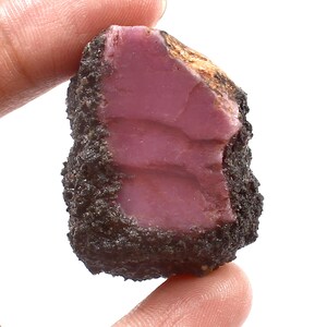 May include: A rough-cut, natural ruby gemstone with a deep pink interior and a dark, textured outer layer. The ruby is held between fingers, showcasing its raw, unpolished beauty. The stone's surface has a slightly uneven texture.