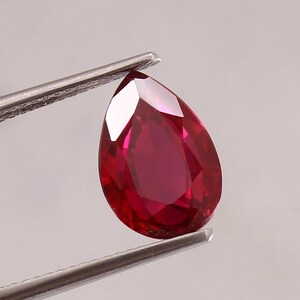 Op de afbeelding: Een gefacetteerde, peervormige robijn edelsteen, vastgehouden door een metalen pincet. De robijn heeft een diepe, verzadigde rode kleur met meerdere reflecterende oppervlakken. De pincet is zilverkleurig en houdt de edelsteen tegen een neutrale achtergrond.