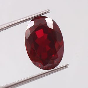 Op de afbeelding: Een dieprode ovale edelsteen met een facetgeslepen vorm. De edelsteen wordt vastgehouden door een pincet.