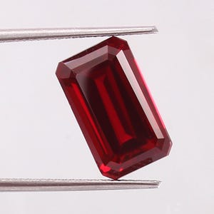 May include: A deep red gemstone with an emerald cut, featuring a rectangular shape and faceted surfaces that reflect light. The stone is held by metal tweezers, showcasing its vibrant colour, clarity, and precise geometric edges in a clear view.