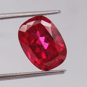 May include: A vibrant, cushion-cut red gemstone held by silver-toned metal tweezers. The stone features a faceted surface that reflects light, creating deep crimson and bright scarlet highlights throughout its transparent, polished crystalline structure.