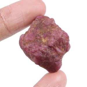 Puede incluir: Una gema de rubí rosa en bruto con un tono rojo oscuro. El rubí tiene una forma irregular y un acabado natural sin pulir.