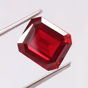 May include: A vibrant, deep red gemstone cut into a rectangular emerald shape with faceted edges. The stone is held by silver-toned metal tweezers, showcasing its clarity and saturated color against a plain, light-colored background.
