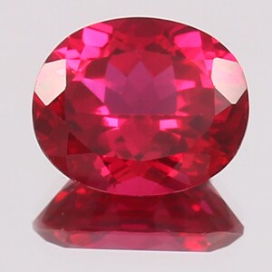 May include: A vibrant, oval-cut gemstone with a deep, saturated red colour and multifaceted surface that reflects light. The stone is positioned above its own clear reflection, showcasing its brilliant clarity and intense, uniform crimson hue.