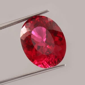 Op de afbeelding: Een gefacetteerde, ovaalvormige robijn edelsteen, vastgehouden door een zilveren pincet. De robijn vertoont een dieprode tint met lichtere roze-rode interne reflecties. De slijping van de edelsteen onthult meerdere facetten.