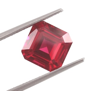 May include: A vibrant, deep red gemstone with a faceted, rectangular emerald cut. The stone is held securely by a pair of silver-toned metal tweezers, showcasing its transparent clarity and sharp, geometric edges against a plain white background.