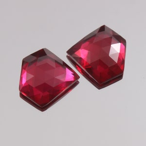 Puede incluir: Dos gemas facetadas en forma de escudo, de un vibrante color rojo rubí. Las gemas tienen un acabado brillante y reflejan la luz, mostrando sus intrincados cortes. Las gemas se muestran sobre una superficie reflectante.