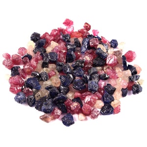 May include: A pile of rough gemstones in various colors, including red, pink, blue, and clear. The gemstones are irregularly shaped and appear to be raw, uncut stones. The image is set against a white background.