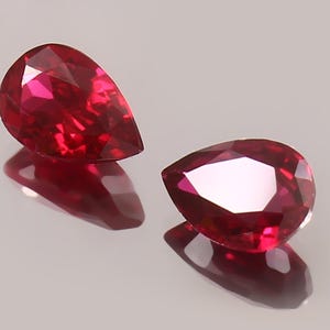 May include: Two pear-shaped ruby gemstones, one on the left and one on the right, are shown on a grey surface. The gemstones are a deep red colour and have a high polish.