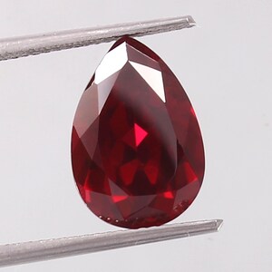 Könnte beinhalten: Ein birnenförmiger, tiefroter Edelstein mit einer facettierten Oberfläche, die das Licht reflektiert. Der Stein wird von einer silberfarbenen Metallpinzette gehalten und zeigt seine transparente, leuchtend karmesinrote Farbe und den präzisen Schliff.