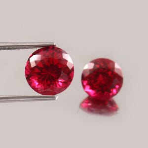 以下が含まれることがあります： 2つの丸いファセットカットが施された鮮やかな赤い宝石。1つは銀色の金属製ピンセットで保持されており、光を反射する複数の三角形のファセットを持つブリリアントカットが特徴です。もう1つの宝石は近くに置かれており、同じ深い赤色をしています。