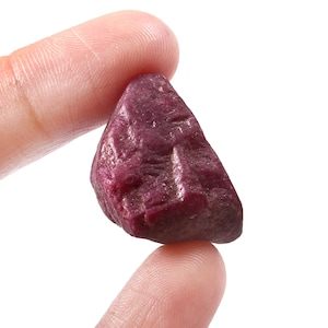 May include: A rough, uncut ruby gemstone, held between two fingers. The ruby is a deep, rich red color with a textured surface. The gemstone is irregularly shaped, showcasing its natural, unpolished state.