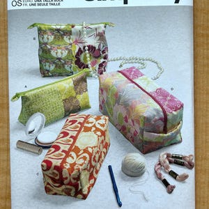 May include: Simplicity S9525 sewing pattern envelope featuring four rectangular zippered pouches in various floral and geometric fabric prints. The image displays finished pouches labeled A, B, C, and D, alongside small accessories like a mirror, yarn, and a crochet hook.