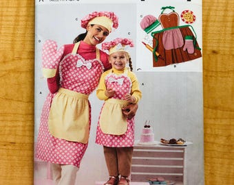 May include: Simplicity sewing pattern S3050 for aprons, hats, and oven mitts. The pattern features a pink polka dot apron with a yellow pocket and bow. The image shows a woman and child wearing the aprons. The text "easy-to-sew" is visible.