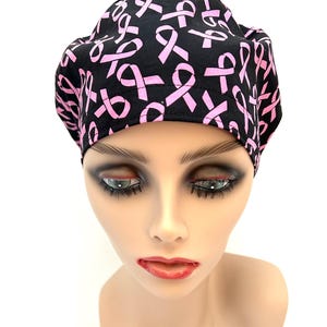 May include: A black scrub cap featuring a repeating pattern of pink awareness ribbons. The fabric head covering has a structured front band and a gathered back section, providing a consistent fit across the top of the head with visible stitching.