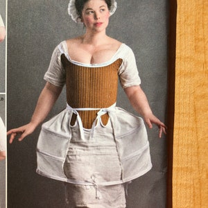 Pu&ograve; includere: Una donna indossa un corsetto bianco e marrone con una gonna bianca e un berretto di pizzo bianco. Il corsetto ha un nastro marrone legato in vita.