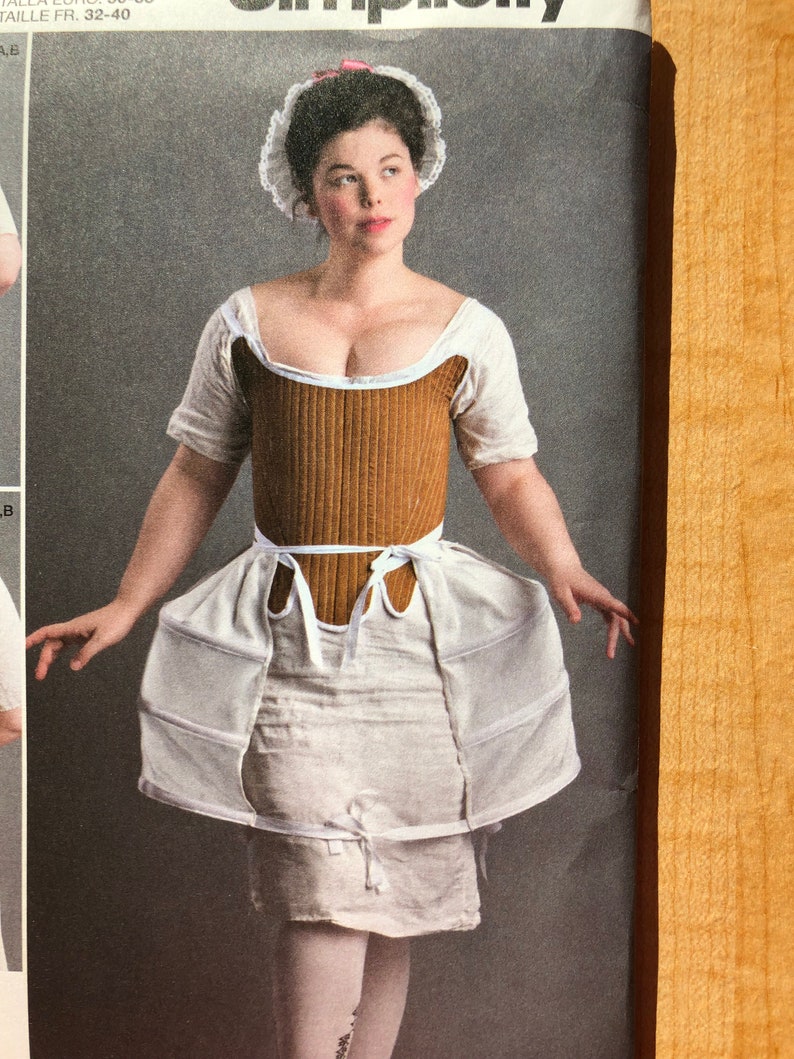 Pu&ograve; includere: Una donna indossa un corsetto bianco e marrone con una gonna bianca e un berretto di pizzo bianco. Il corsetto ha un nastro marrone legato in vita.