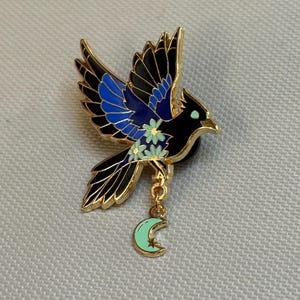 May include: A gold-toned metal enamel pin shaped like a bird in flight with black and blue wings, decorated with small light green flowers. A small, dangling light green crescent moon charm is attached to the bird by a gold-toned metal chain.