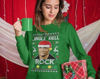 May include: A woman wearing a green long-sleeve Christmas sweater with the text "JINGLE BELL ROCK" and a graphic of a person wearing a Santa hat. She holds a green mug and a wrapped gift with a red and green plaid pattern.