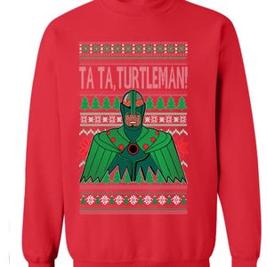 May include: Red crewneck sweatshirt featuring a graphic of a superhero with a green costume and a brain-shaped helmet. The text "TA TA, TURTLEMAN!" is above the superhero. The design includes Christmas tree and snowflake patterns.