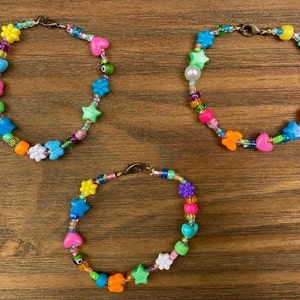 May include: Three colorful beaded bracelets with various shapes and colors, including stars, hearts, flowers, and a pearl. The bracelets are made with small plastic beads and are strung on a silver chain.