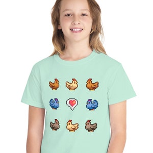 Pu&ograve; includere: Una maglietta verde chiaro con un design a pixel di galline e un cuore. Le galline sono di diversi colori, tra cui marrone, giallo e blu.