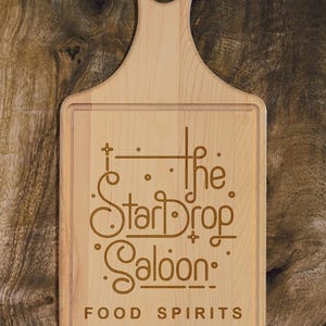 May include: A light-colored wooden cutting board with a handle and a rectangular cutting surface. The board features the engraved text "The StarDrop Saloon" with the words "FOOD SPIRITS" below. The board is set against a wooden background.