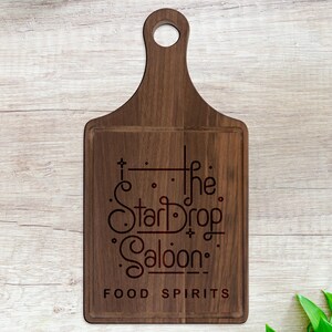 May include: A wooden cutting board with a handle and the engraved text "The StarDrop Saloon: FOOD SPIRITS." The board is rectangular with a rounded top and a hole for hanging. The wood has a natural grain.
