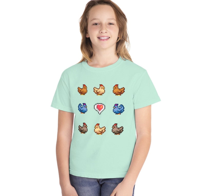 Pu&ograve; includere: Una maglietta verde chiaro con un design a pixel di galline e un cuore. Le galline sono di diversi colori, tra cui marrone, giallo e blu.