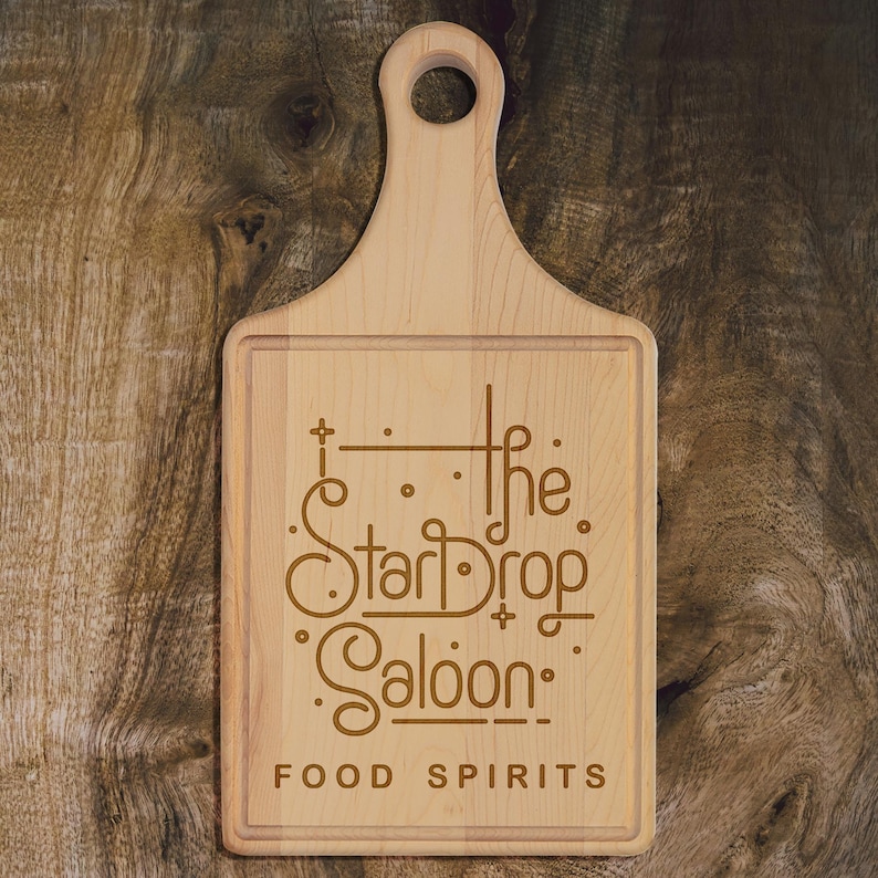 May include: A light-colored wooden cutting board with a handle and a rectangular cutting surface. The board features the engraved text "The StarDrop Saloon" with the words "FOOD SPIRITS" below. The board is set against a wooden background.