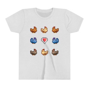 Pu&ograve; includere: Una maglietta bianca con un design a pixel di galline e un cuore. Le galline sono marroni, gialle e blu.