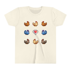 Pu&ograve; includere: Una maglietta beige con un design a pixel di galline e un cuore. Le galline sono marroni, gialle e blu.