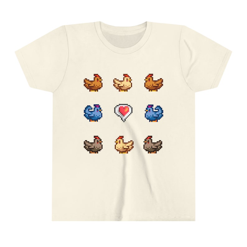 Pu&ograve; includere: Una maglietta beige con un design a pixel di galline e un cuore. Le galline sono marroni, gialle e blu.