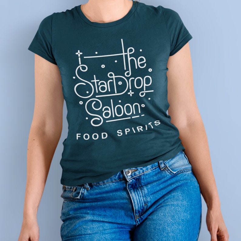 May include: A teal t-shirt with the text "The StarDrop Saloon: Food Spirits" in white, stylized lettering. The shirt is paired with blue denim jeans. The design features a celestial theme, suggesting a bar or restaurant.