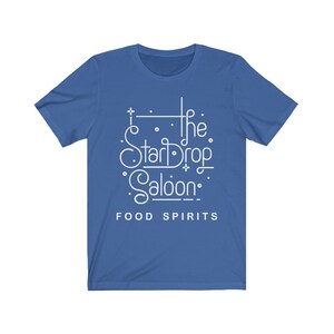 May include: Royal blue t-shirt with white text that reads "The StarDrop Saloon" in a stylized font, with the words "FOOD SPIRITS" below. The shirt has short sleeves and a crew neck.
