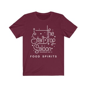 May include: A maroon t-shirt with the white text "The StarDrop Saloon" in a stylized font, with the words "FOOD SPIRITS" below. The shirt has short sleeves and a crew neck.