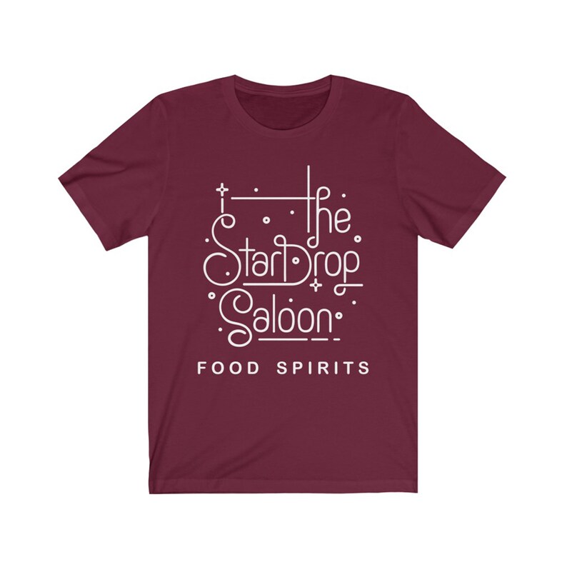 May include: A maroon t-shirt with the white text "The StarDrop Saloon" in a stylized font, with the words "FOOD SPIRITS" below. The shirt has short sleeves and a crew neck.