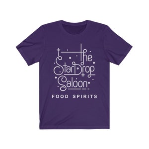 May include: A purple t-shirt with the white text "The StarDrop Saloon" in a stylized font. Below the name are the words "FOOD SPIRITS" in a simple sans-serif font. The shirt has short sleeves and a crew neck.