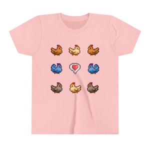 Pu&ograve; includere: Una maglietta rosa con un design a pixel di galline e un cuore. Le galline sono marroni, gialle e blu.